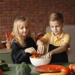 Cheerful girl helping little boy in apron preparing salad while standing in kitchen near table with mix of fresh vegetables behind brick wall