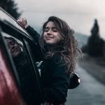 Contented young female with brown hair fluttering in wind leaning out of window of moving car in countryside
